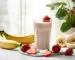 Batido de frutas con leche en un vaso, rodeado de fresas, banana y mango.